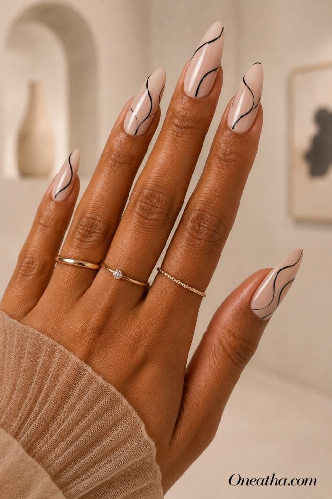 Luxury nail designs by Oneatha featuring glossy nude almond nails with thin black abstract line art accents, delicate gold rings, and a soft beige editorial background with framed silhouette artwork.