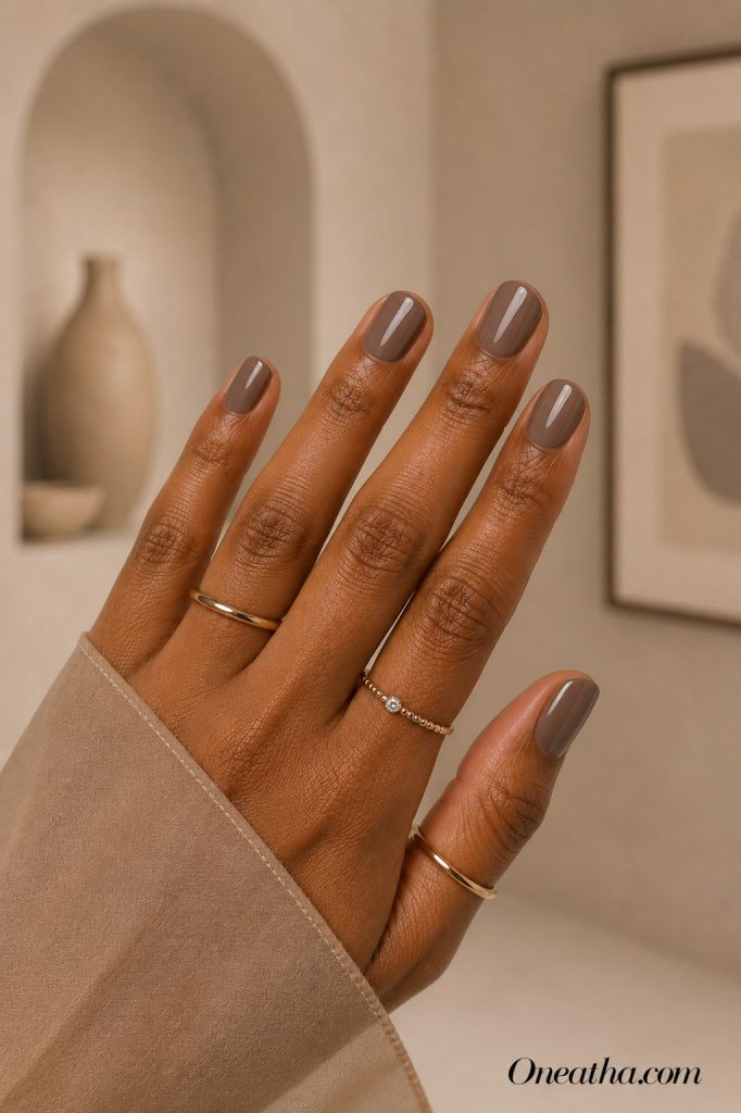 Luxury nail designs by Oneatha featuring glossy muted taupe short rounded nails, minimalist neutral manicure, gold rings, and a warm beige editorial background.