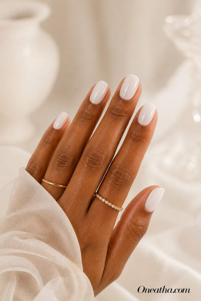 Luxury milky white nail designs by Oneatha featuring glossy almond nails with an ultra-gloss finish, gold rings, and a soft ivory editorial background with dreamy bridal-inspired lighting.