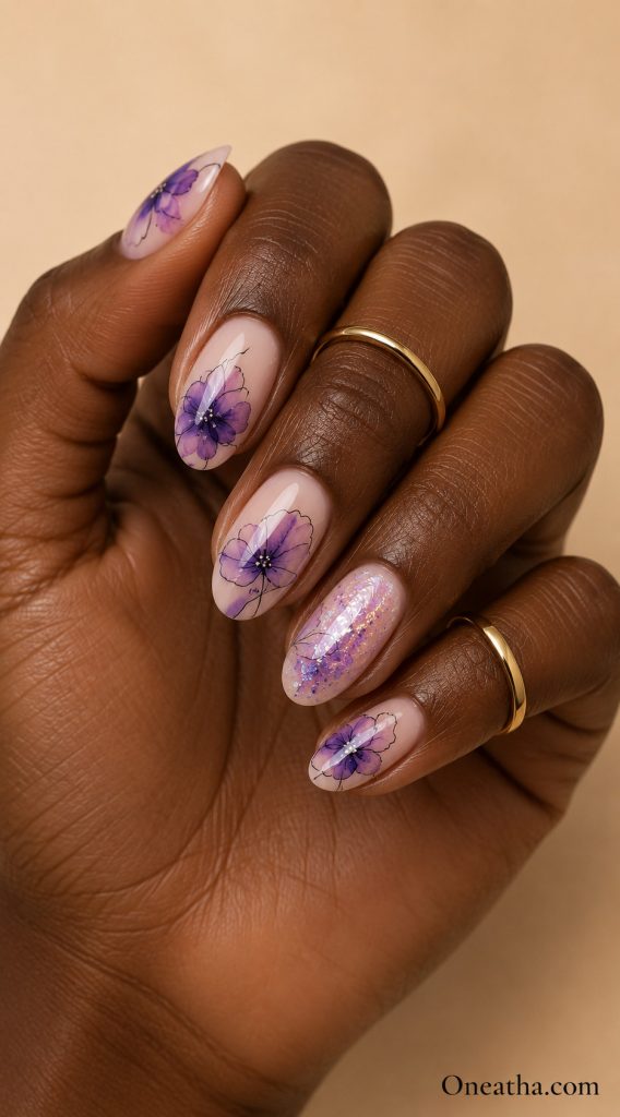 Glossy sheer nude almond nails by Oneatha featuring delicate purple inked floral artwork and a sparkling lilac glitter accent nail on a warm brown hand model with minimalist gold rings.
