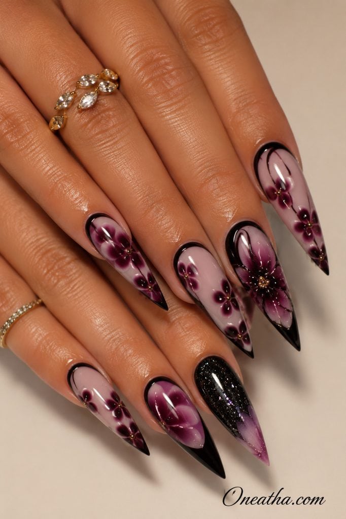 Glossy stiletto nails by Oneatha featuring deep purple floral nail art, black French tips, a soft lilac base, and a black shimmer accent nail fading into a light purple aura tip, styled with delicate gold rings.