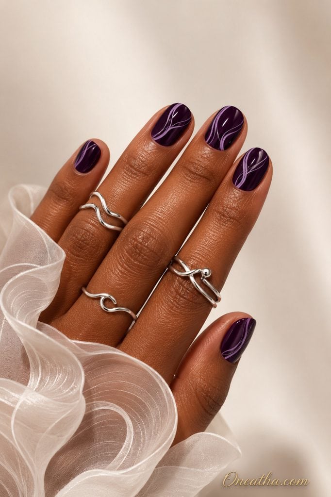 Short glossy plum nails by Oneatha featuring delicate lilac swirl line art, styled with sculptural silver rings and a sheer white organza sleeve against a soft luxury neutral background.