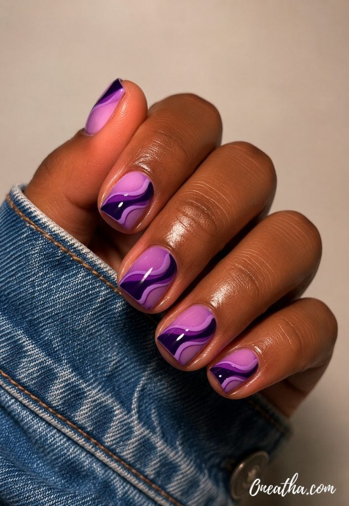 Short glossy nails by Oneatha featuring abstract lilac, violet, and deep purple swirl nail art with a high-shine finish, styled with a denim jacket sleeve against a soft neutral background.