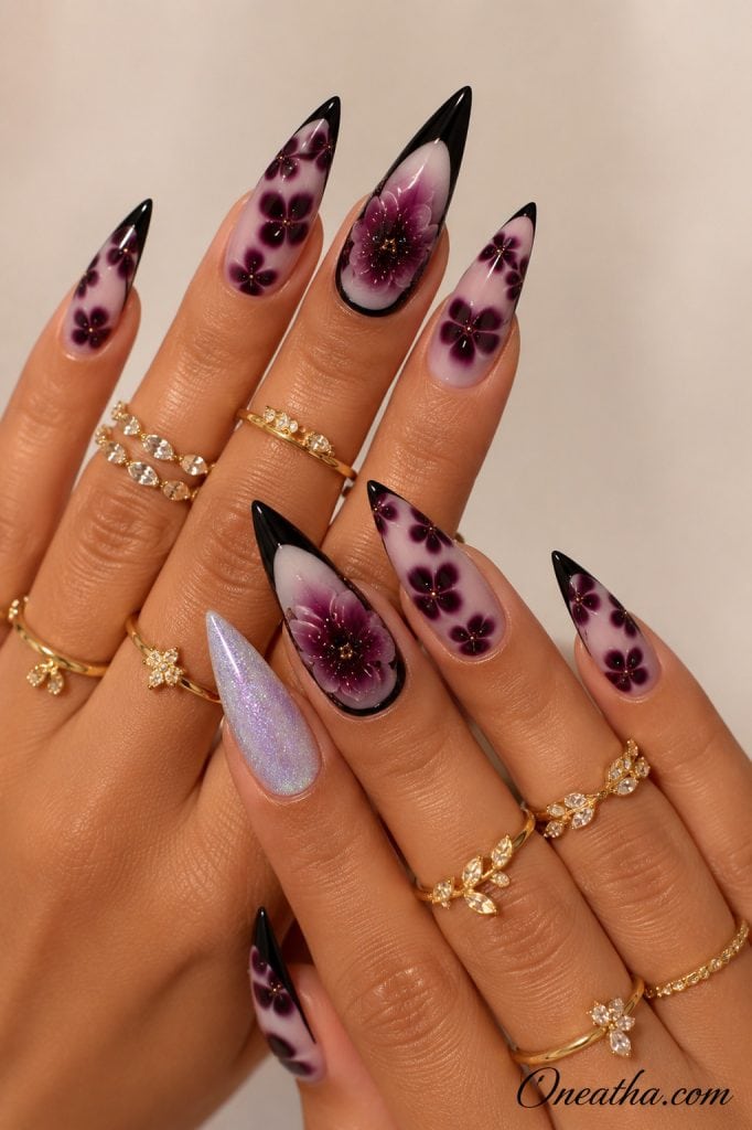 Glossy stiletto nails by Oneatha featuring deep plum floral nail art, black French tips, a shimmering lavender glitter accent nail, and delicate gold rings, creating a romantic midnight-inspired manicure against a soft neutral background.
