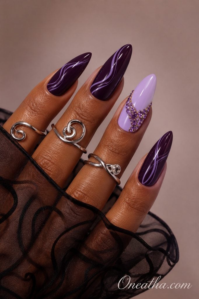 Glossy almond nails by Oneatha featuring deep amethyst purple polish with delicate lilac swirl line art, a soft lavender accent nail embellished with sparkling amethyst crystals, sculptural silver rings, and a sheer black organza sleeve against a luxury neutral background.