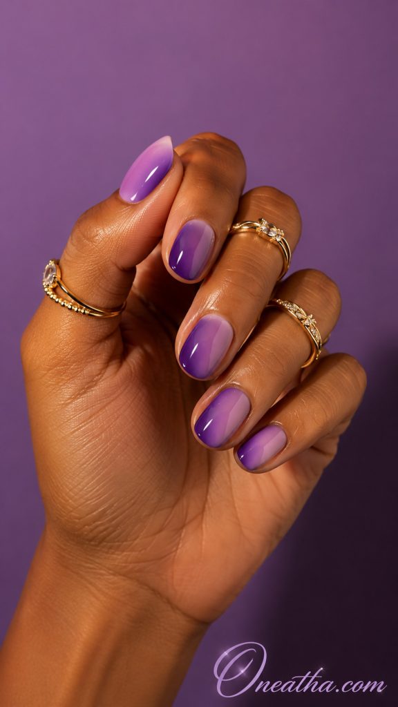 Glossy purple and lilac ombré short square nail design by Oneatha on a warm cinnamon-toned hand model wearing luxe gold rings against a plain purple background.