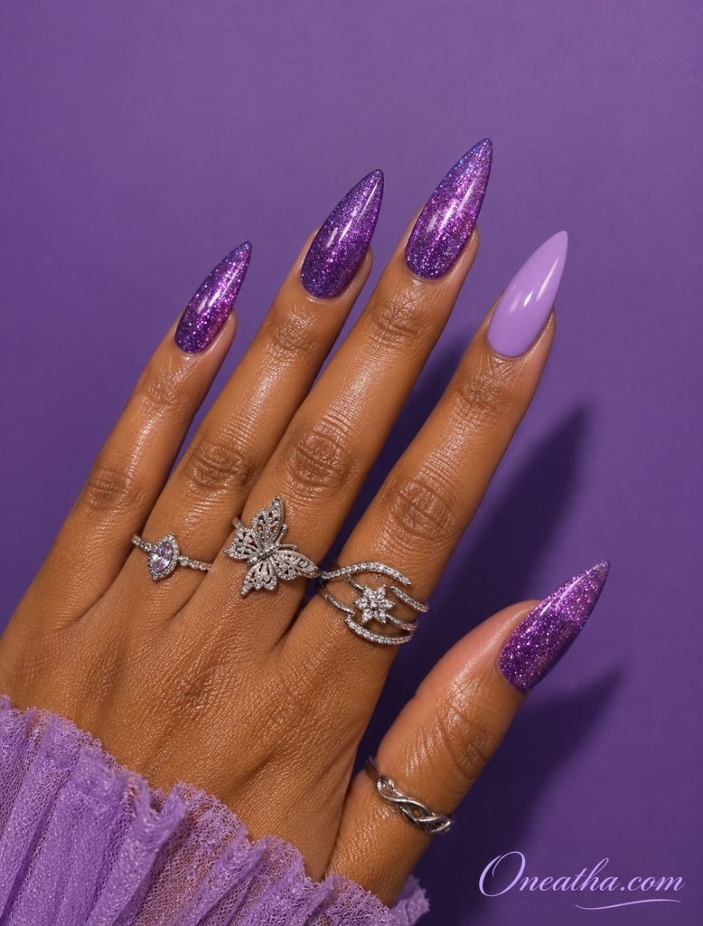 Glossy purple glitter nails by Oneatha featuring a soft lilac accent nail, stilleto-shaped manicure, luxe silver rings, and a rich purple background.