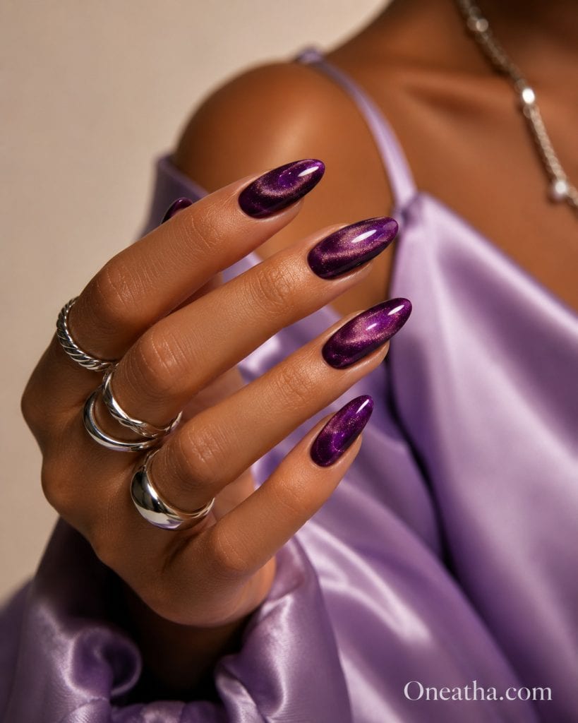 Glossy purple cat-eye nails by Oneatha featuring almond-shaped nails, a shimmering magnetic finish, silver rings, and a lilac satin blouse in a luxury editorial beauty style.
