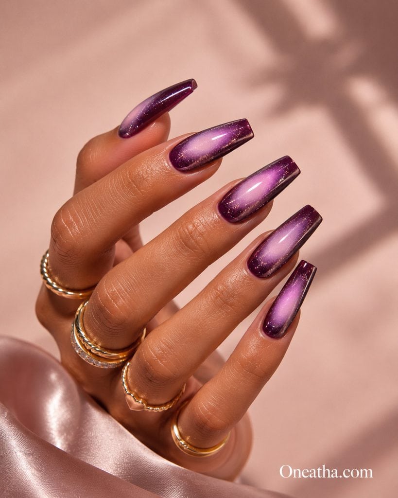 Glossy purple cat-eye nails by Oneatha featuring a luminous violet gradient, long coffin-shaped nails, gold rings, and a soft blush editorial background with a luxurious beauty aesthetic.